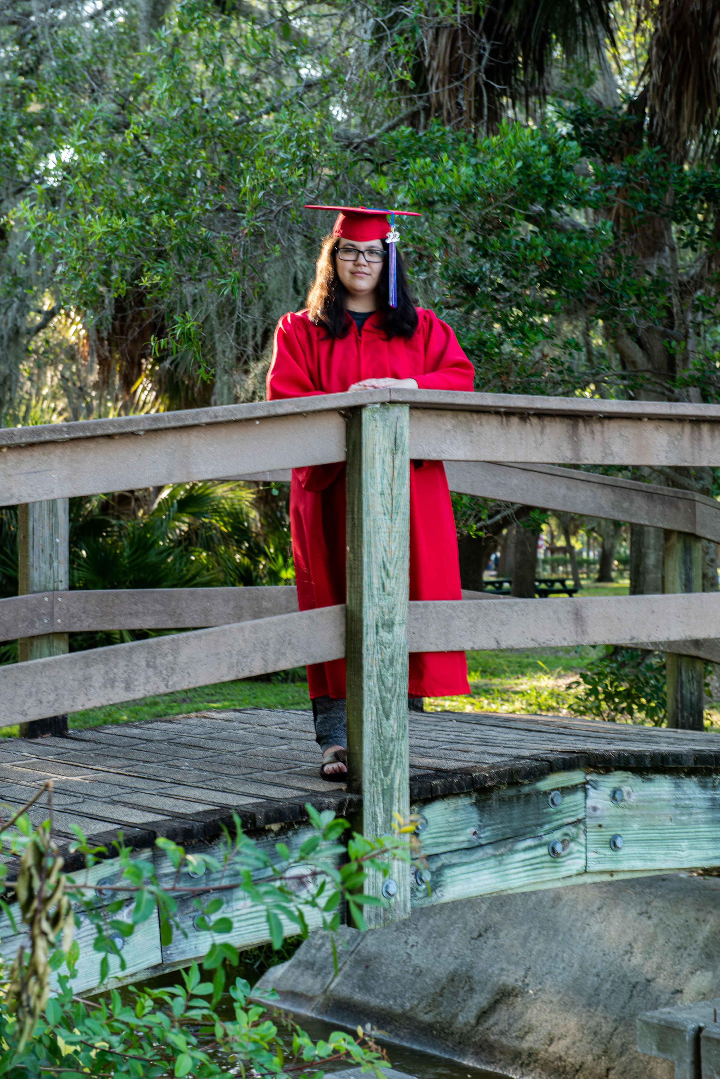 Senior in red graduation cap and gown leaning on wooden bridge railing in Florida park